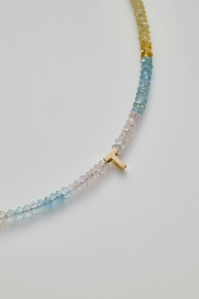 A close-up image of the One Letter Bracelet in Aquamarine and 18k Gold, featuring a small 18k solid gold 'T' charm. 14K gold letter P necklace strung with naturally shaded aquamarine gemstones, showcasing ocean-inspired hues.