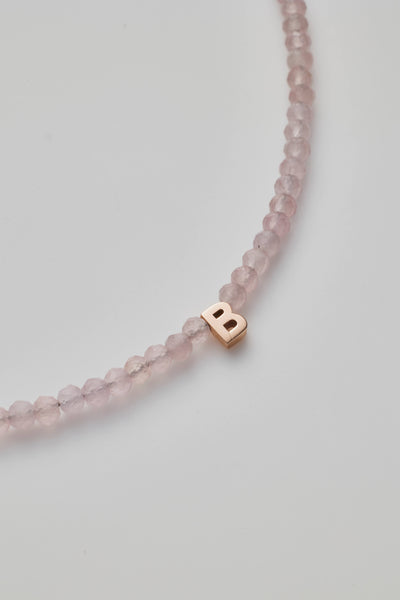 The One Letter Bracelet in Rose Quartz and 14k Rose Gold features small, round rose quartz beads and a gold lobster clasp. A single 14k gold initial charm, the letter "B," is threaded onto the beads. The bracelet is available in custom sizes, arranged in a simple, curved shape on a white background.