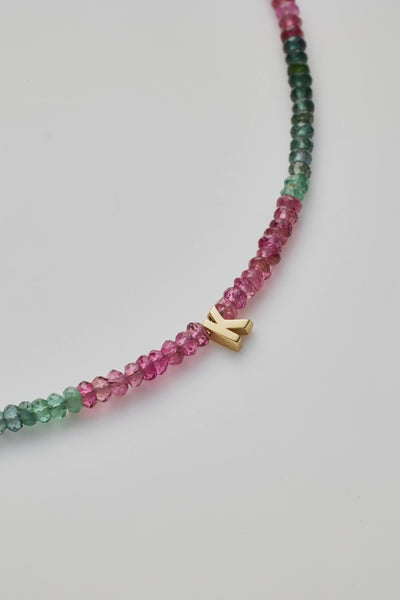 The One Letter Bracelet in Tourmaline and 14k Gold showcases delicate, small multicoloured beads arranged in a beautiful gradient from pink to green. The piece is elegantly finished with an 14k solid gold clasp and a charming letter "K" charm. Displayed on a white background in an oval shape, the bracelet is available in custom sizes.