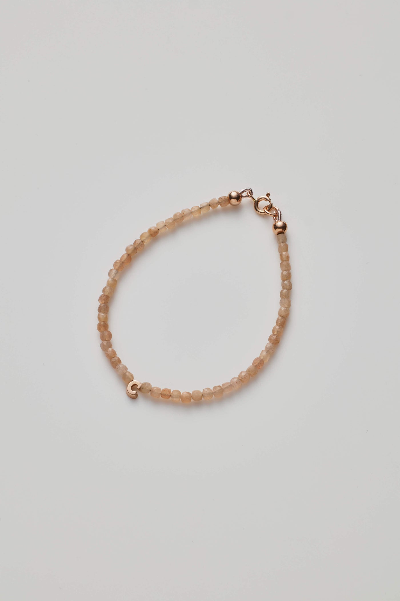 The One Letter Bracelet in Blush Moonstone and 14k Gold is a delicate, custom-sized piece featuring small, round blush moonstone beads evenly spaced along its length. The light beige hues of the moonstones add a soft elegance, while the 14-karat gold lobster clasp and findings provide a subtle yet refined touch to its simple design.