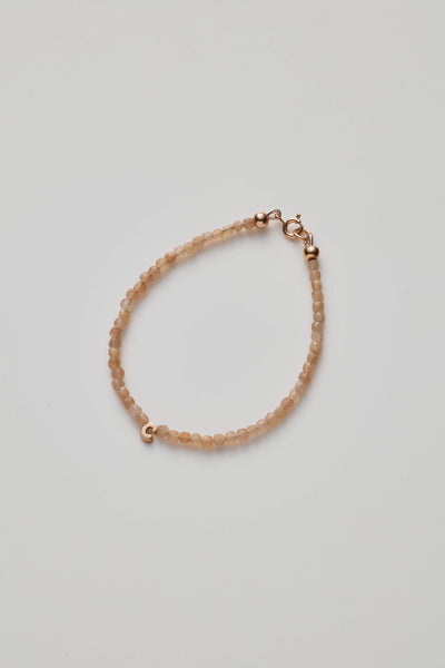 The One Letter Bracelet in Blush Moonstone and 14k Gold is a delicate, custom-sized piece featuring small, round blush moonstone beads evenly spaced along its length. The light beige hues of the moonstones add a soft elegance, while the 14-karat gold lobster clasp and findings provide a subtle yet refined touch to its simple design.