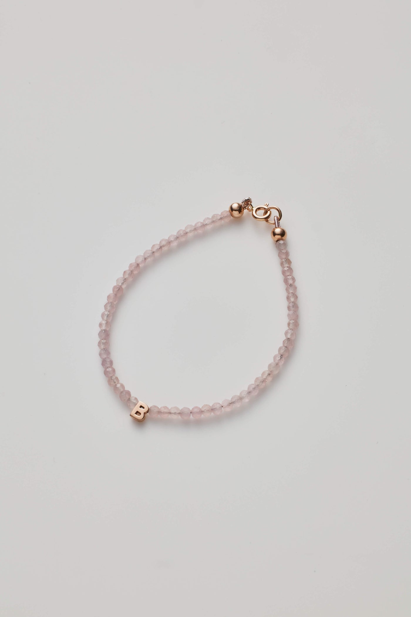 The One Letter Bracelet in Rose Quartz and 14k Rose Gold features small, round rose quartz beads and a gold lobster clasp. A single 14k gold initial charm, the letter "B," is threaded onto the beads. The bracelet is available in custom sizes, arranged in a simple, curved shape on a white background.