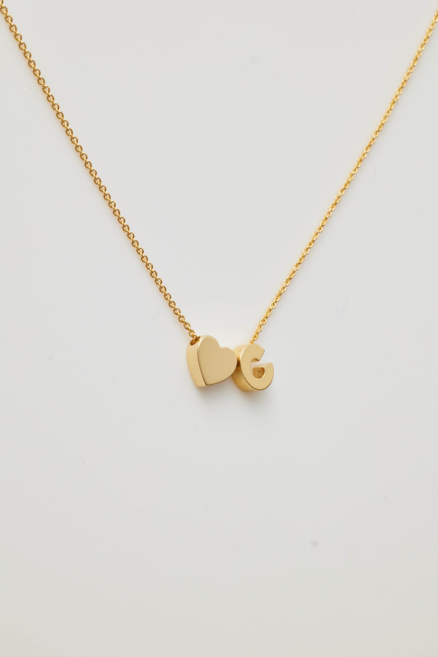 The One Letter and Heart Necklace in 18k Gold features a delicate solid gold chain adorned with two pendants: a heart and a "C". The heart and letter contribute to a simple, elegant design against a white background, exuding timeless charm.