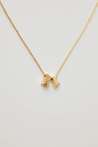 The Two Letters Necklace in Diamonds and 18k Gold showcases a minimalist and elegant design with two hanging pendants: a solid 18k gold "K" and a gold "K" encrusted with sparkling diamonds. This stunning necklace shines beautifully against a grey background.