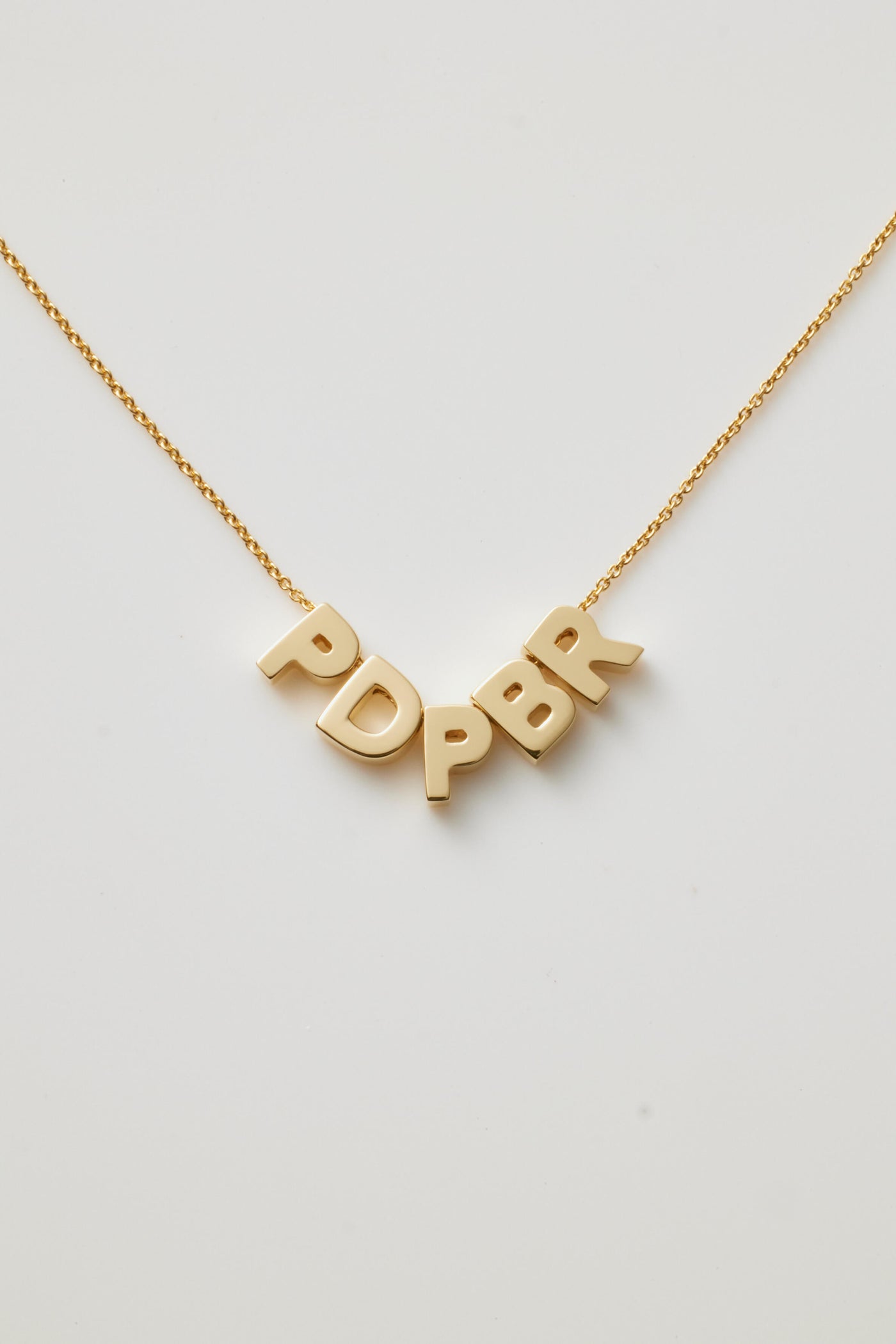 The Five Letters Necklace in 18k Gold is a delicate chain necklace adorned with five solid gold letters - PDPBR hanging gracefully at the centre. These evenly spaced letters form an abstract or acronymic design that stands out beautifully against a white background.