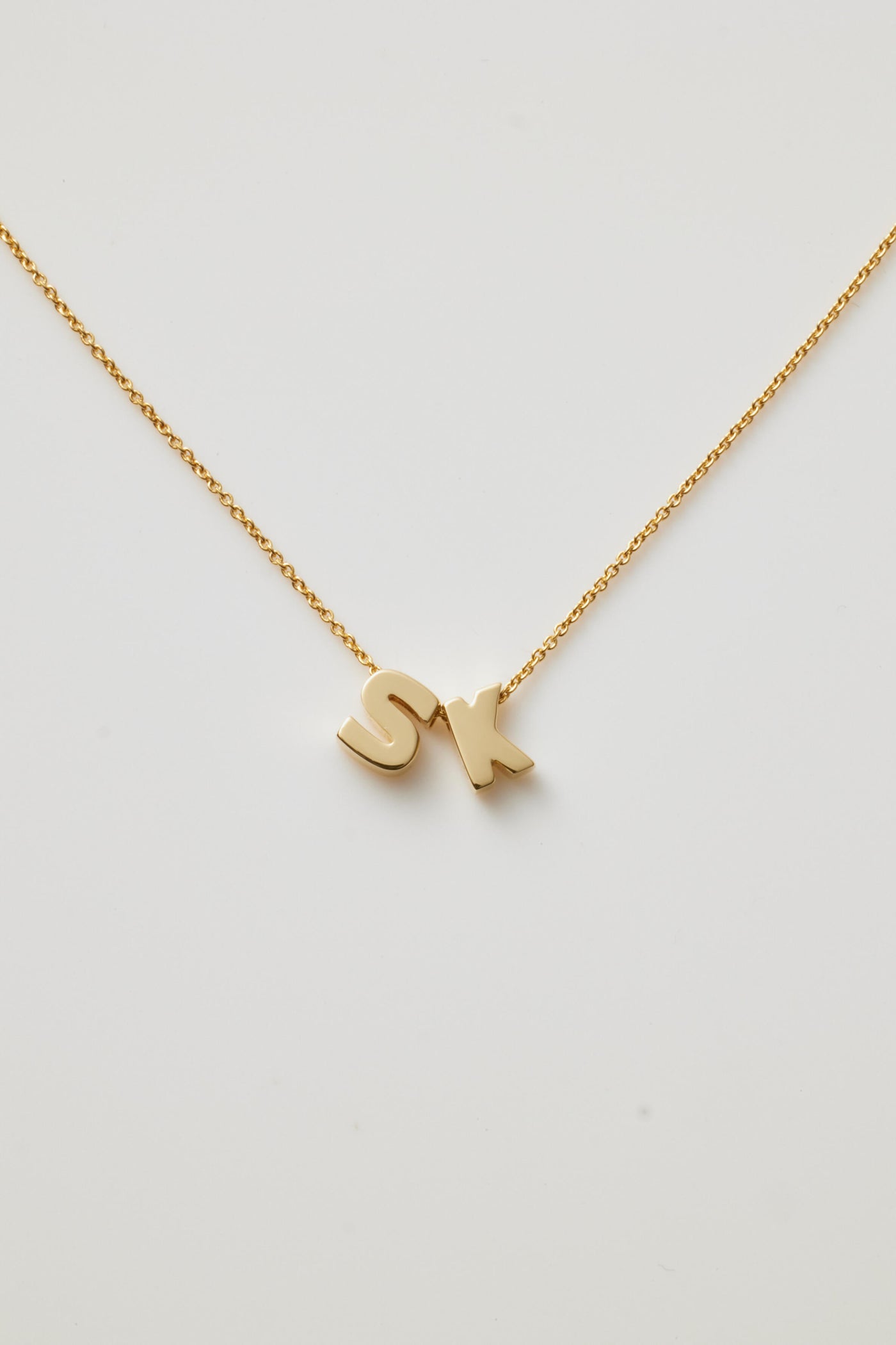 The Two Letters Necklace in 18k Gold showcases a delicate 18k solid gold chain with two gold letter charms, "S" and "K," elegantly positioned close together at the centre, all set against a plain white background.