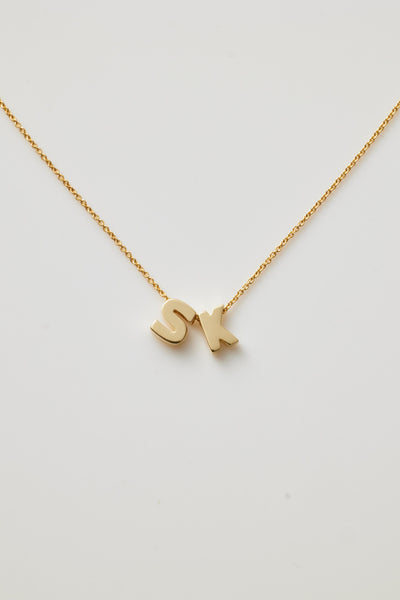 The Two Letters Necklace in 18k Gold showcases a delicate 18k solid gold chain with two gold letter charms, "S" and "K," elegantly positioned close together at the centre, all set against a plain white background.