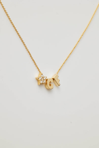 The "Two Letters and Diamond Star Necklace in 18k Gold" is a delicate piece showcasing two letter charms, "G" and "F," flanking a radiant star-shaped charm adorned with round brilliant diamonds. All are elegantly suspended from a fine gold chain against a white background.