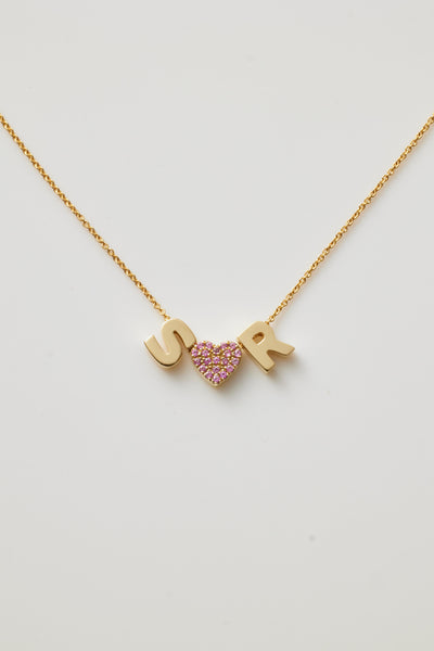 A delicate Two Letters and Pink Sapphire Heart Necklace in 18k Gold features three hanging pendants with two gold letters "SR." and "heart "the heart is set with vibrant hued pink sapphires, encrusted in gemstones. The necklace lies gracefully on a white background.