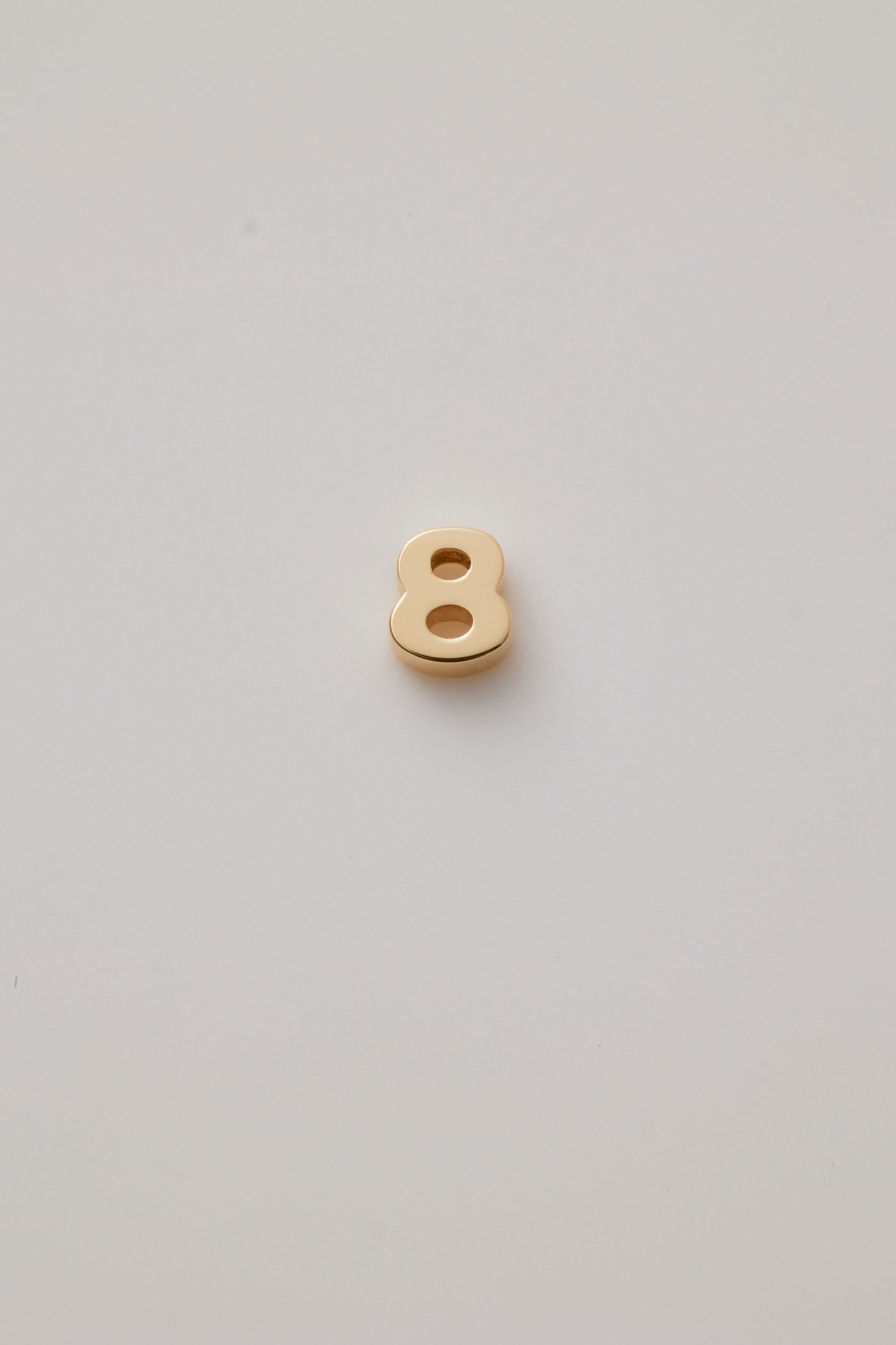 Three-dimensional number charms, each representing a number from 0 to 9, crafted from 18-karat solid gold. Each charm has a sleek, polished surface that radiates a rich, golden glow, adding a touch of luxury. A small hole is integrated at the top of each charm for easy attachment to a chain or bracelet. 