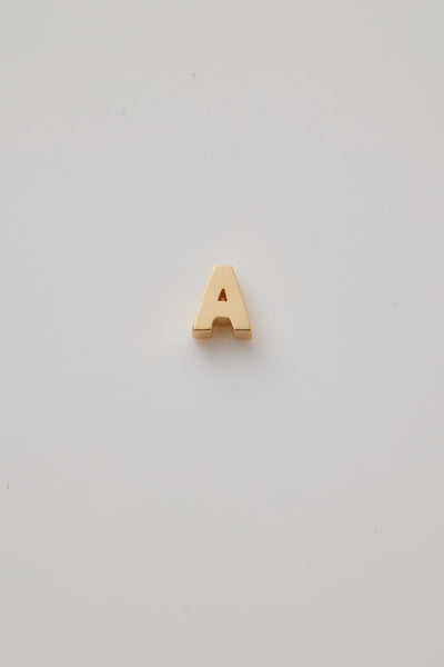 A three-dimensional charm crafted from 18-karat gold, shaped into a bold and elegant letter from the alphabet. The charm has a smooth, polished surface that gleams under light, highlighting its luxurious golden hue. Its edges are slightly rounded for a refined and sophisticated finish. The charm features a small, discreet hole at the top, allowing it to be easily attached to a chain or bracelet. The overall design is both modern and timeless, exuding a sense of opulence and personalisation.