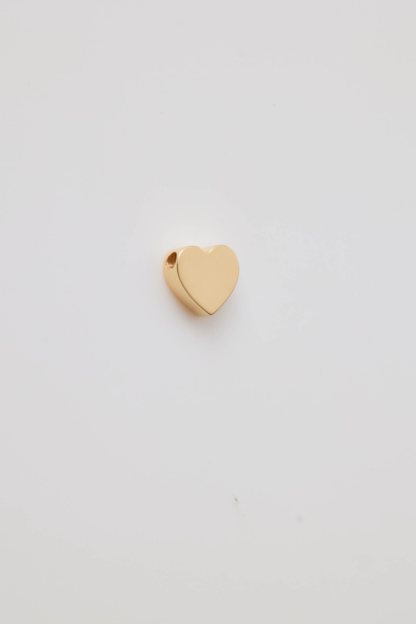 Gold heart-shaped pendant with a smooth, polished finish, shown from an angled side view. A small hole for threading a chain is visible near the top edge, emphasising its minimalist and elegant design. The pendant is set against a plain white background