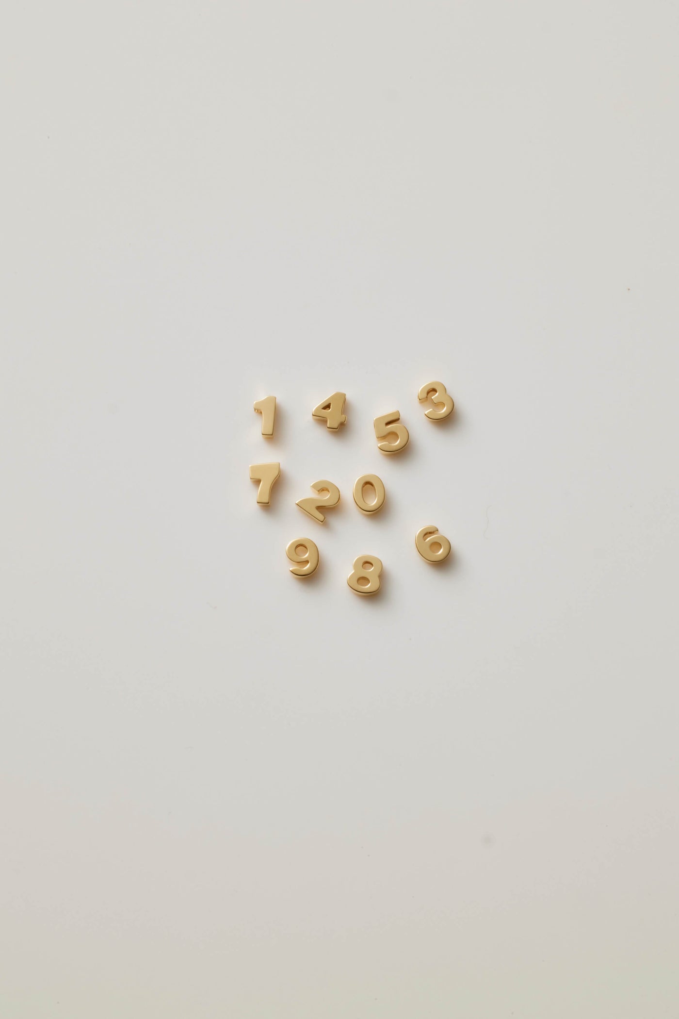 Three-dimensional charms, each representing a number from 0 to 9, crafted from 18-karat solid gold. Each charm has a sleek, polished surface that radiates a rich, golden glow, adding a touch of luxury. The numbers are bold and slightly rounded at the edges, ensuring a smooth and refined appearance. A small hole is integrated at the top of each charm for easy attachment to a chain or bracelet. The collection symbolizes luck and individuality, offering a timeless and elegant way to celebrate mean