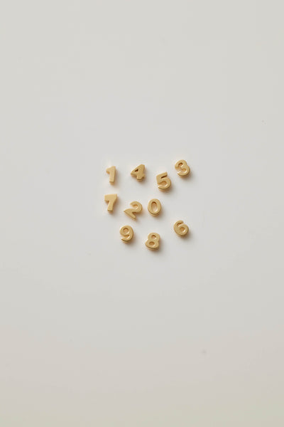 Three-dimensional charms, each representing a number from 0 to 9, crafted from 18-karat solid gold. Each charm has a sleek, polished surface that radiates a rich, golden glow, adding a touch of luxury. The numbers are bold and slightly rounded at the edges, ensuring a smooth and refined appearance. A small hole is integrated at the top of each charm for easy attachment to a chain or bracelet. The collection symbolizes luck and individuality, offering a timeless and elegant way to celebrate mean