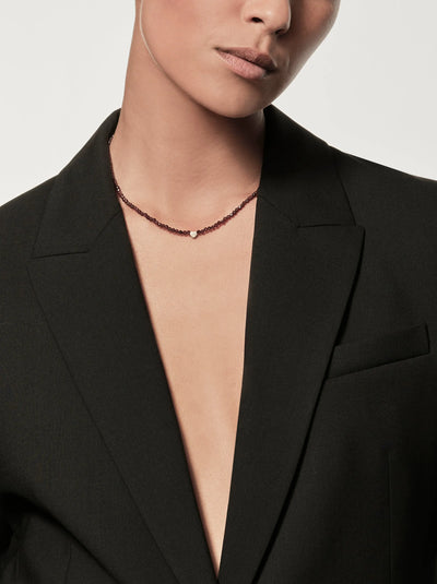 A person is shown from the neck down, donning a deep V-neck blazer over a bare chest. They're accessorized with the Heart Necklace in Garnet and 18k Gold, adding an elegant touch to their ensemble. The plain light background accentuates the minimalistic and sophisticated fashion style.