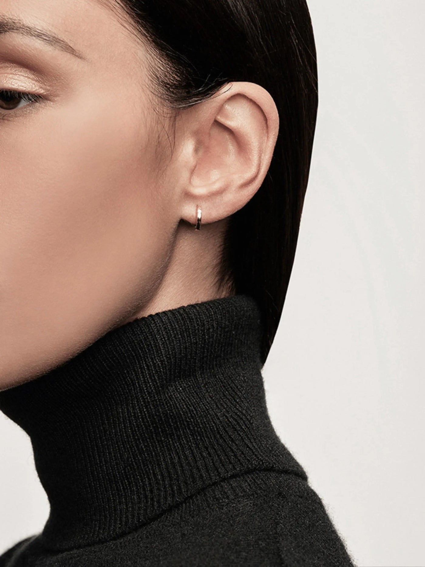 Close-up of a person wearing Huggie Earrings in 18k Gold in their left ear. They have short, dark hair and are wearing a black turtleneck sweater. The background is plain white.