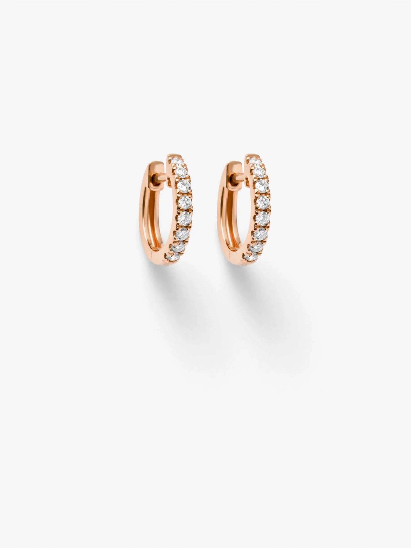 The Huggie Earrings in Diamonds and 18k Rose Gold are elegantly showcased against a plain white background. Each earring, encrusted with a row of clear, brilliant-cut diamonds along the front edge, is positioned side by side, casting subtle shadows below.