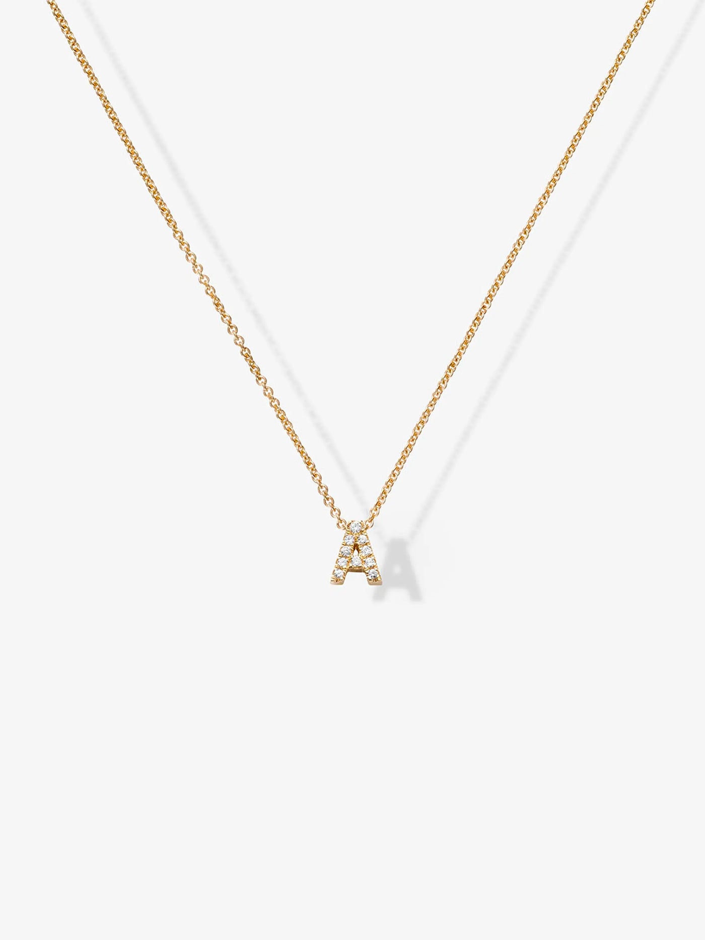 The Letter A Necklace in Diamonds and 18k Gold showcases a delicate pendant shaped like the letter "A," adorned with small, sparkling diamonds. Displayed against a plain, light background, the exquisite pendant hangs gracefully at the center of the image.

