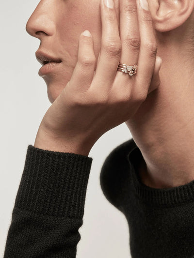 Close-up of a person's face and hand. The person is wearing a black sweater and appears pensive, with their chin resting on their hand adorned with several Letter A Rings featuring round brilliant diamonds set in 18k solid gold. The background is plain and light-colored, drawing focus to the person and jewelry.