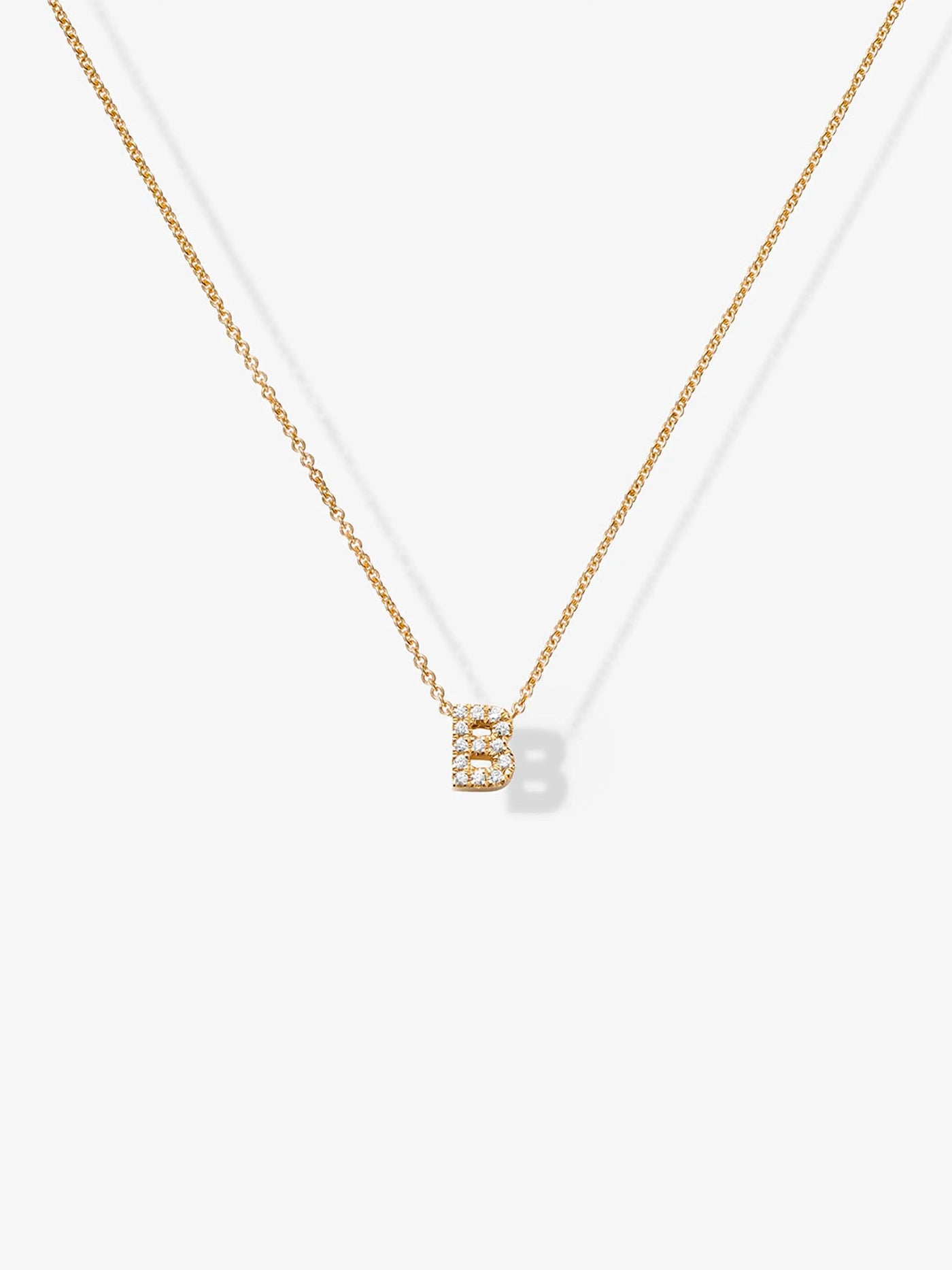 The "Letter B Necklace in Diamonds and 18k Gold" features a delicate 18k gold chain and a customizable diamond letter pendant shaped like "B," encrusted with small, sparkling diamonds. The necklace is set against a plain white background.