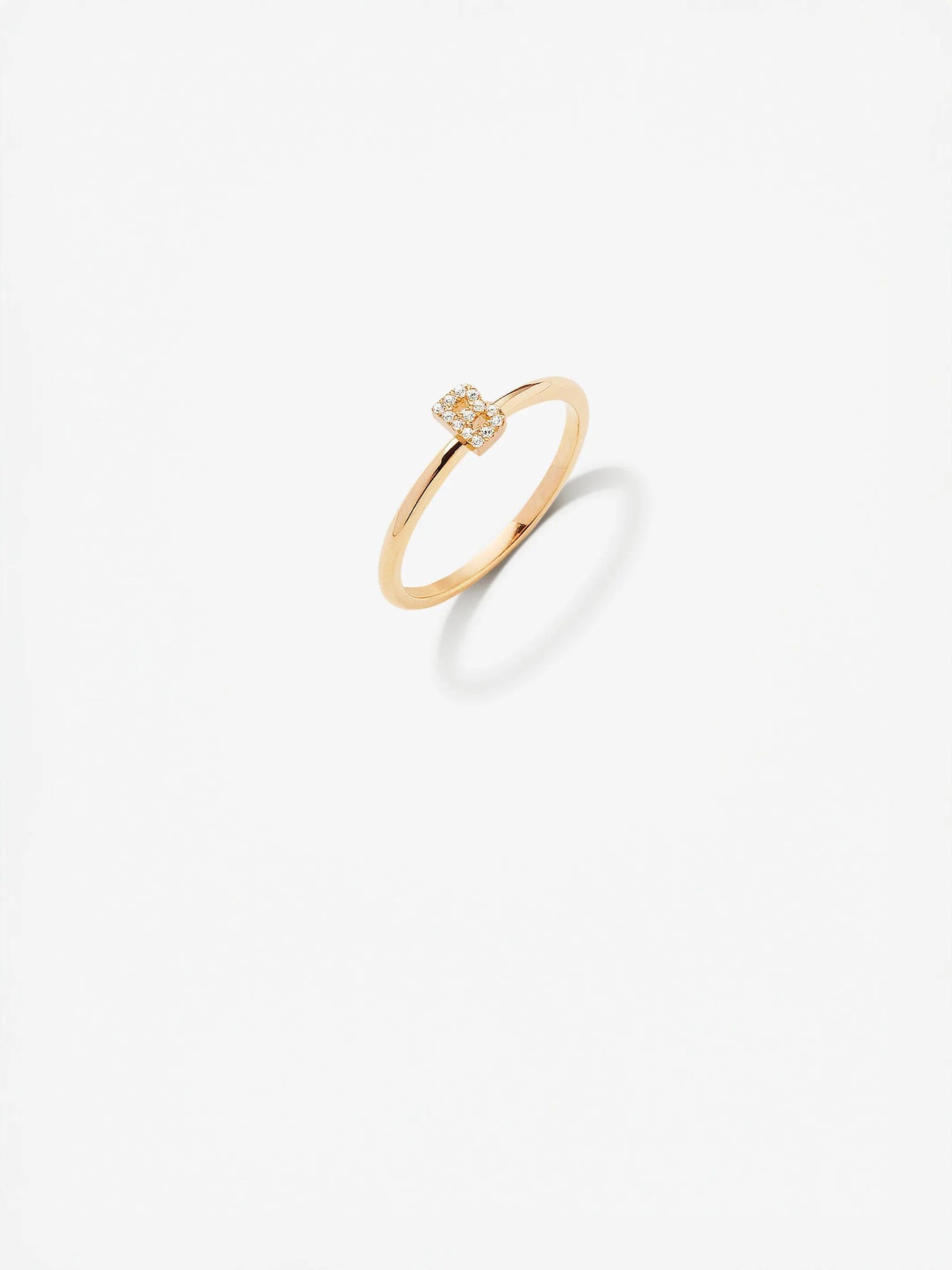 An elegant custom-made Letter B Ring in Diamonds and 18k Gold with a square-shaped setting encrusted with small diamonds, showcased against a minimalistic white background. The ring's shadow is visible slightly to the right on the surface.