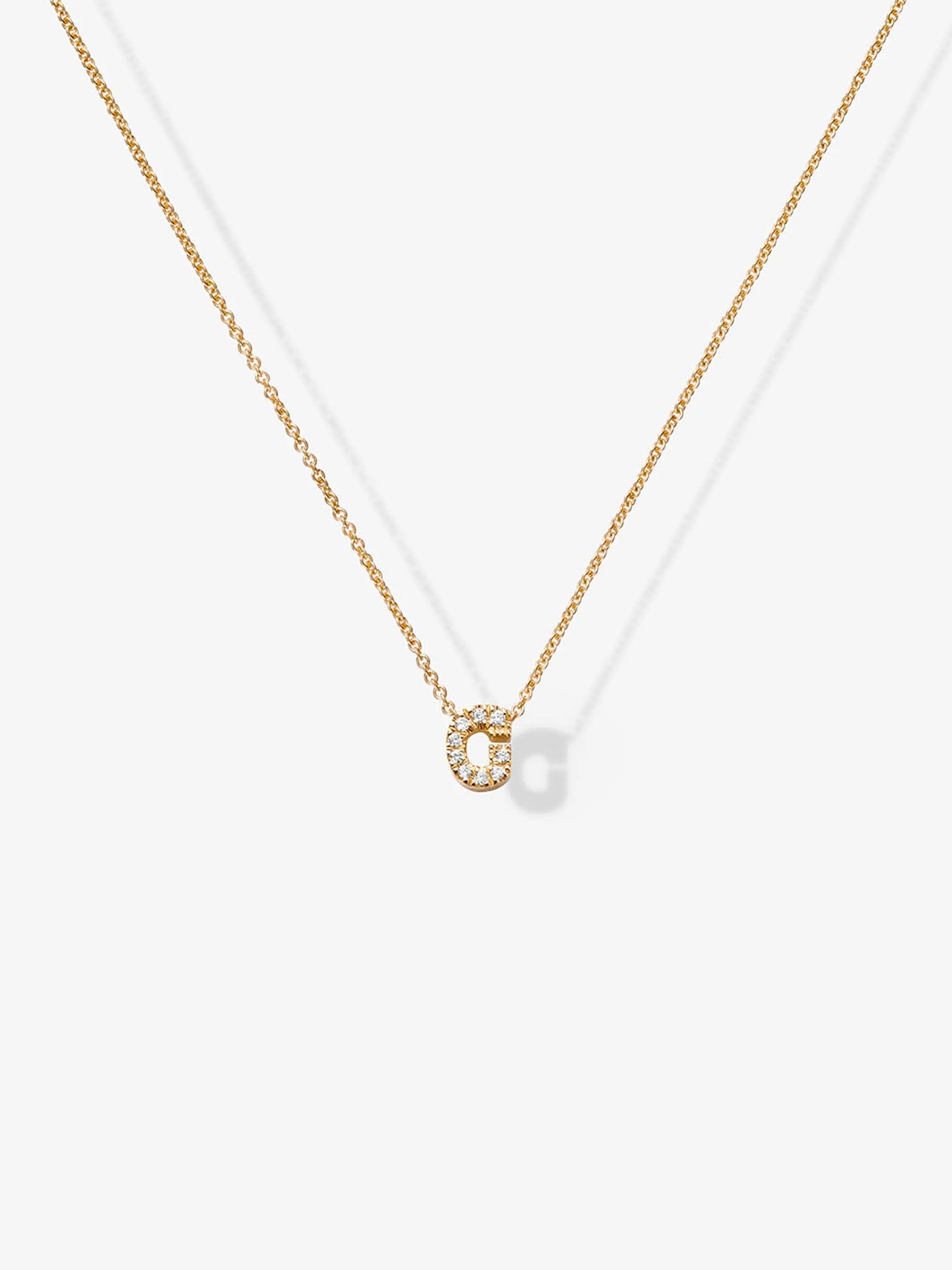 Experience the elegance of our Letter G Necklace in Diamonds and 18k Gold. This delicate necklace features a small, glittering "G" pendant encrusted with tiny, sparkling crystals, adding a touch of sophistication to its minimalist design. The image is set against a plain, light background. Customizable charms make this piece uniquely yours.