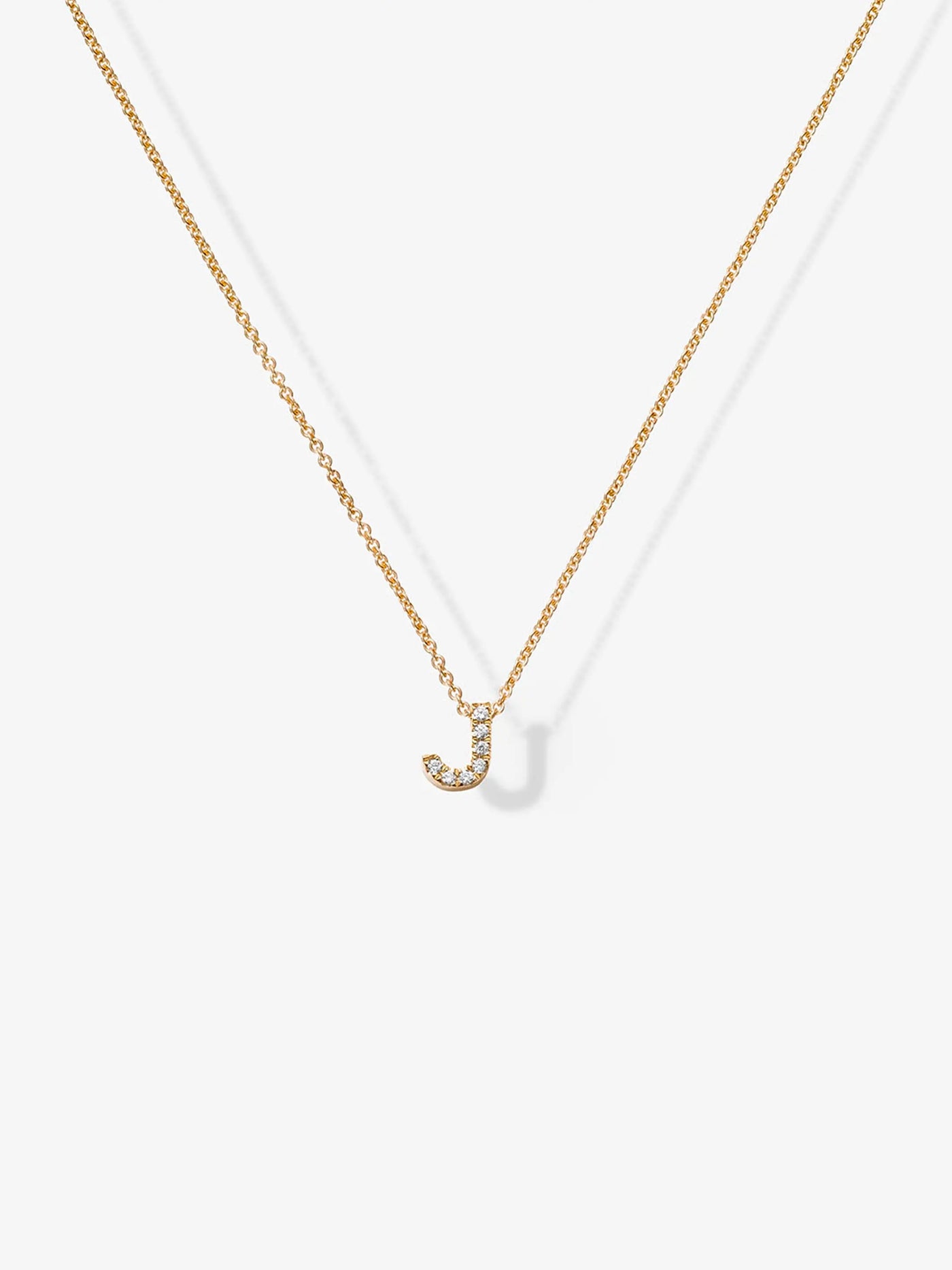 The "Letter J Necklace in Diamonds and 18k Gold" showcases a delicate 18k gold chain with a pendant shaped like the letter "J", adorned with small, sparkling diamonds. The elegant letter necklace hangs at the center, casting a subtle shadow on the plain light background.