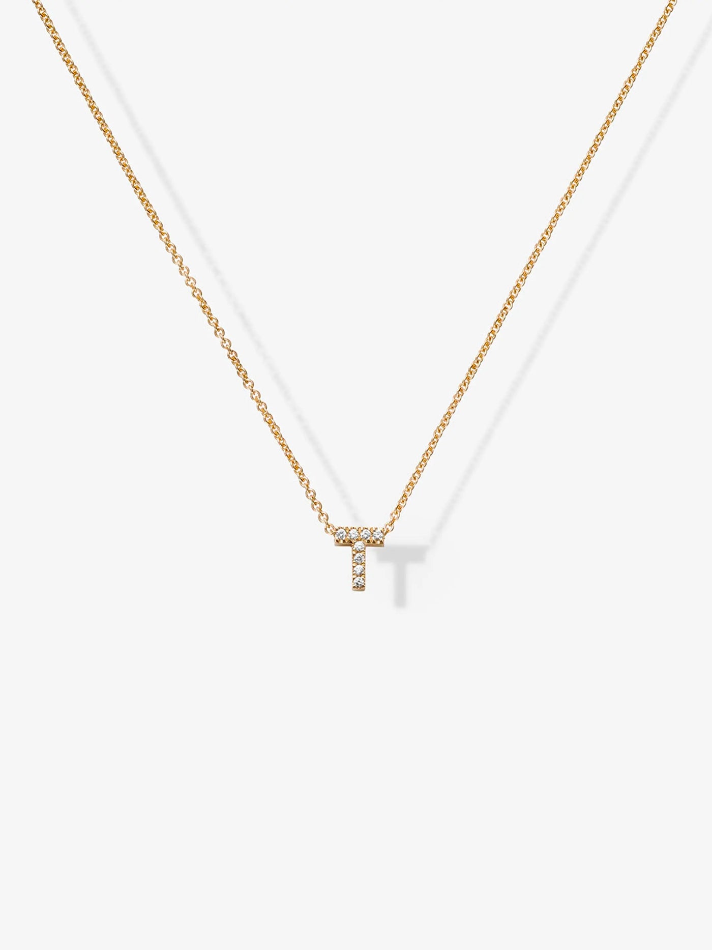 The Letter T Necklace in Diamonds and 18k Gold features a delicate 18k gold chain with a pendant in the shape of the letter "T," adorned with small, sparkling diamonds and set against a plain white background.