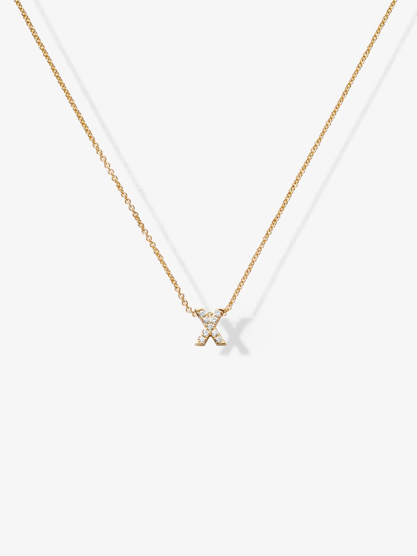 A delicate 18k solid gold necklace with a thin chain features a small, gold "X" pendant encrusted with tiny, sparkling diamonds. This elegant accessory, known as the Letter X Necklace in Diamonds and 18k Gold, is displayed against a plain white background, casting a faint shadow.