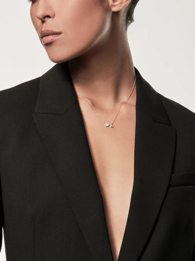 A person wearing an open black blazer and a delicate One Heart and Letter Necklace in Diamonds and 18k Gold, posing against a plain background. Their head is slightly turned to the side, with short hair styled neatly. The image emphasizes style and elegance.