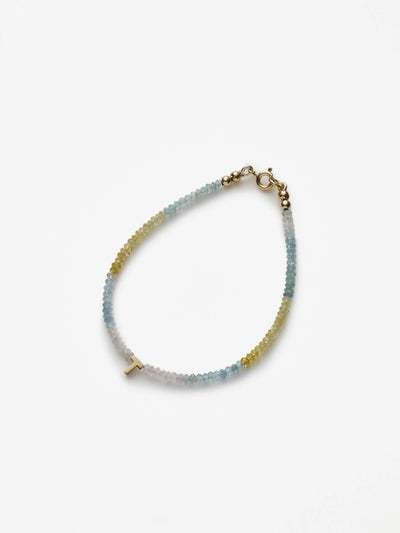 The One Letter Bracelet in Aquamarine and 18k Gold is a delicate piece featuring small, multicolored beads in shades of blue, green, and white. It includes a tiny 18k solid gold clasp and an additional small gold cross charm attached. Ideal for mental clarity, the bracelet is displayed on a plain white background.