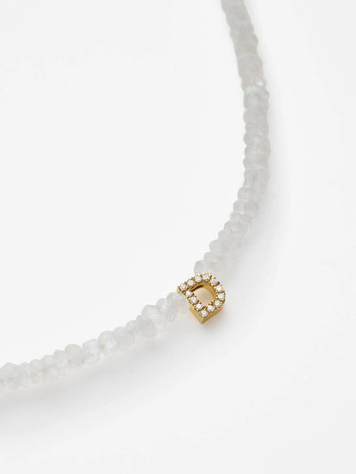 A close-up of the One Letter Bracelet in Diamonds, Rainbow Moonstone and 18k Gold reveals a strand of rough, translucent moonstones. The bracelet features an 18k solid gold charm adorned with tiny white diamonds. The charm is slightly off-center on the string of moonstones against a plain white background.