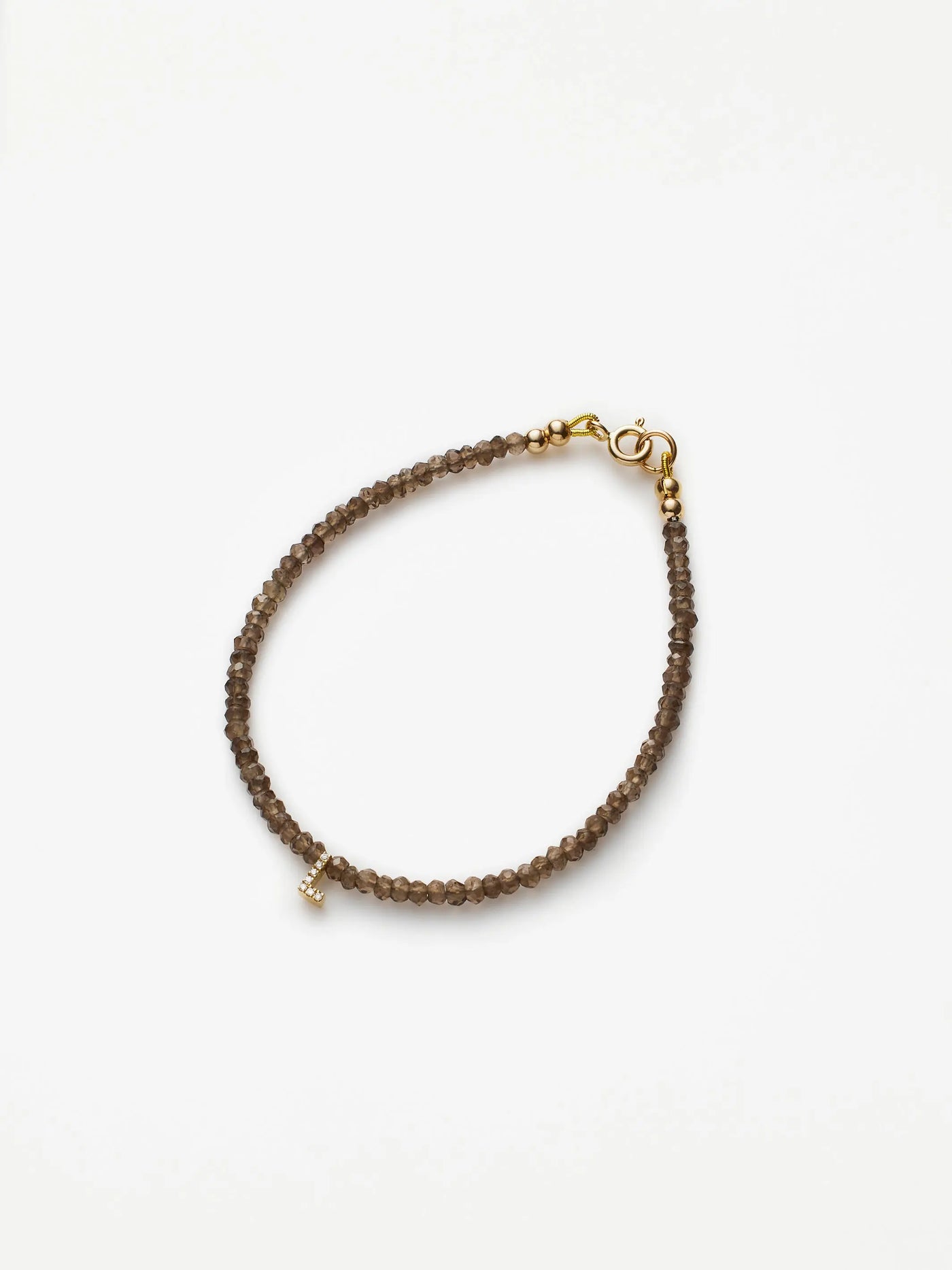 The One Letter Bracelet in Diamonds, Smoky Quartz and 18k Gold is a delicate, hand-strung piece featuring small, smoky brown quartz beads connected by a string. Accentuated with a tiny gold-colored charm shaped like a lowercase "t" near the clasp and finished with an 18k solid gold lobster clasp, this elegant bracelet is displayed on a white background.