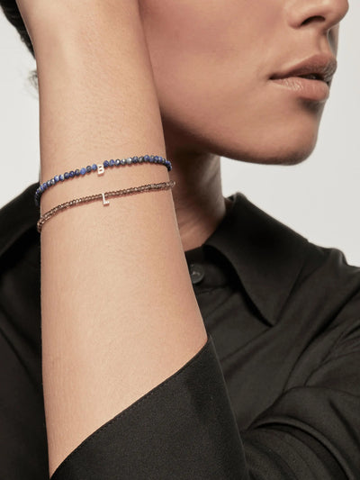 A close-up of a person with their hand resting on their head, showcasing two delicate beaded bracelets on the wrist. One bracelet is an 18k solid gold piece featuring blue beads with a tiny charm; the other is the One Letter Bracelet in Diamonds, Smoky Quartz, and 18k Gold with brown beads and a similar charm. The person is wearing a black shirt.