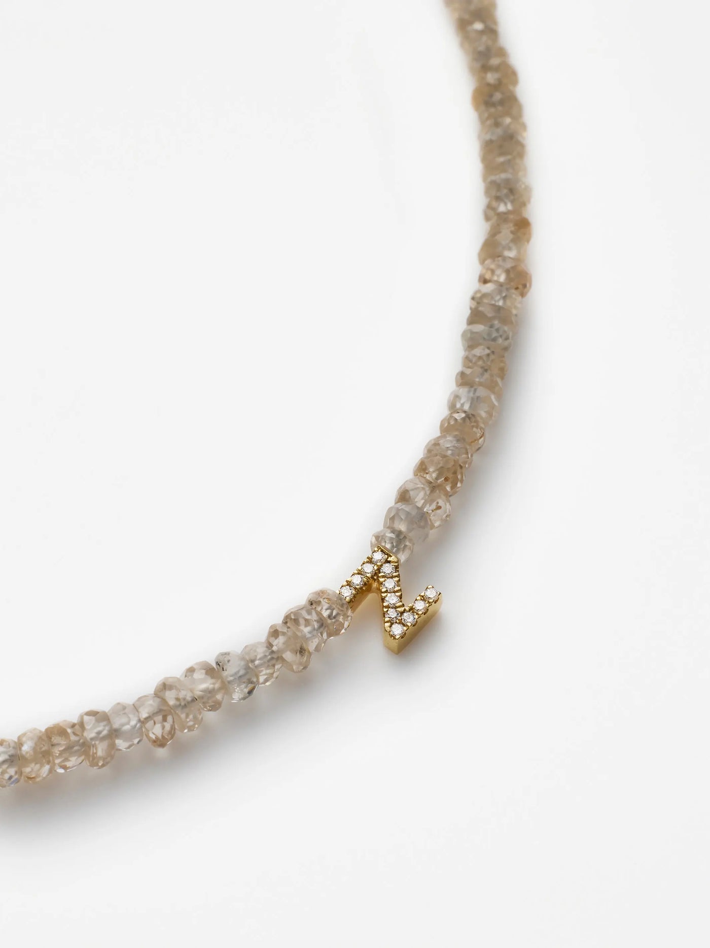 A close-up image of the One Letter Bracelet in Diamonds, Zircon and 18k Gold featuring light-colored, faceted beads. At the center of the bracelet is a small, 18k gold letter "N" pendant encrusted with tiny clear crystals. The piece is displayed against a white background.