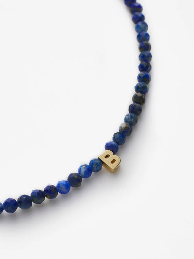 A close-up of the One Letter Bracelet in Lapis Lazuli and 18k Gold, showcasing small, round, faceted blue beads. At its center is a single 18k solid gold bead shaped like the letter "B." The bracelet is photographed against a plain white background.