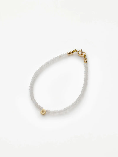 The One Letter Bracelet in Rainbow Moonstone and 18k Gold features small, translucent beads with a single 18k solid gold bead displaying the letter "D." This elegant bracelet includes a gold clasp closure and can be customized to fit different sizes. The product is showcased against a plain white background.