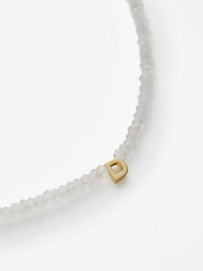 A close-up image of the One Letter Bracelet in Rainbow Moonstone and 18k Gold, showcasing its translucent white stones. This customizable bracelet features a single 18k solid gold letter "D" charm threaded onto it. The background is a plain white surface.