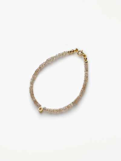 Introducing the One Letter Bracelet in Zircon and 18k Gold: a delicate bracelet adorned with a thin strand of small, translucent zircon beads. A single 18k solid gold bead adds a touch of sophistication, and the bracelet is secured with a gold clasp. The simple yet elegant design stands out beautifully against a plain, light background. Custom sizes are available upon request.