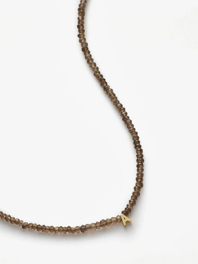 A delicate One Letter Necklace in Smoky Quartz and 18k Gold, made of small, round, translucent smoky quartz beads arranged in a single strand. The necklace features a small gold letter "A" charm near the bottom and an 18k gold chain. The white background emphasizes the simplicity and elegance of the jewelry.