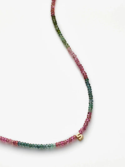 The One Letter Necklace in Tourmaline and 18k Gold showcases a delicate design with small, multicolored tourmaline gemstones in green, pink, and red shades. It includes a slightly off-center, small 18k gold 'S' charm. The beads are threaded on a fine, near-invisible string for a minimalist appearance.