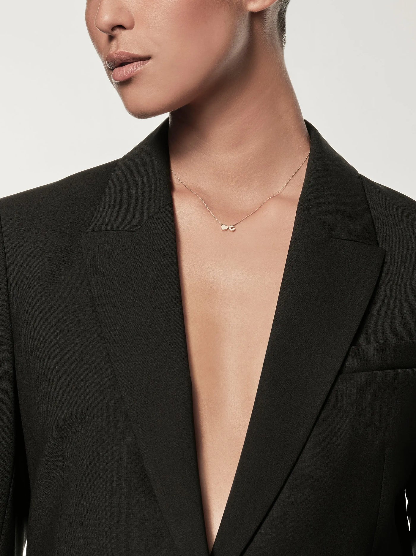 A person with short hair is wearing the One Letter and Heart Necklace in 18k Gold from versejewellery.com and a stylish black blazer with a deep V-neckline. Their head is turned to the side against a plain white background.