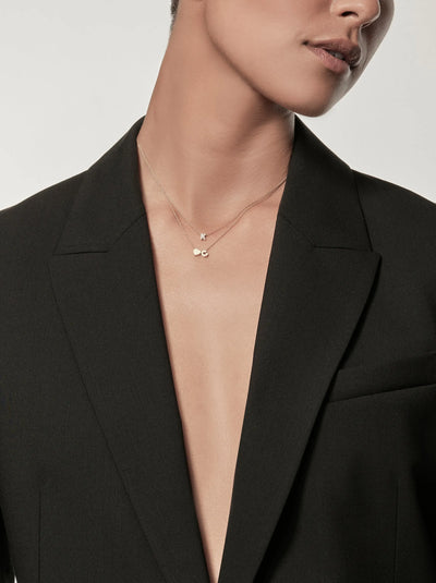 The image features a person from the shoulders to the top of the head, with their eyes and forehead excluded. They are wearing a black blazer with a deep neckline and an 18k solid gold necklace from versejewellery.com, showcasing small letters "K" and "C". The background is light-colored.