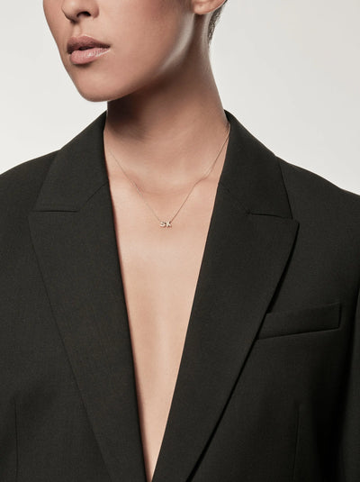 A person wearing a black blazer with no shirt underneath, showcasing a delicate Two Letters Necklace in 18k Gold with the initials "SK" at the center. The background is a plain light gray. The person's face is mostly out of frame, focusing on the jewelry and attire.