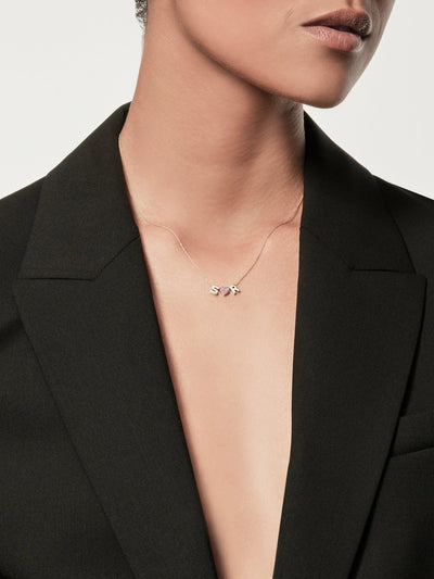 The Two Letters and Ruby Heart Necklace in 18k Gold is a delicate piece, showcasing letter charms "S" and "R" flanking a small ruby heart adorned with pink gemstones. The heart is elegantly positioned between the two letters, all displayed against a plain white background.